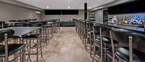 Uma área de bar moderna com um balcão comprido e vários bancos. Há mesas e cadeiras dispostas ao fundo.