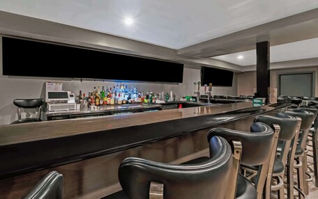 A bar area with a long counter, multiple bar stools, a television mounted on the wall, and a refrigerator.