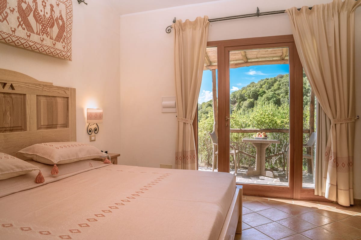 A bedroom with a large bed, a wooden headboard, a nightstand with a lamp, and a view of a balcony with a table and chairs.