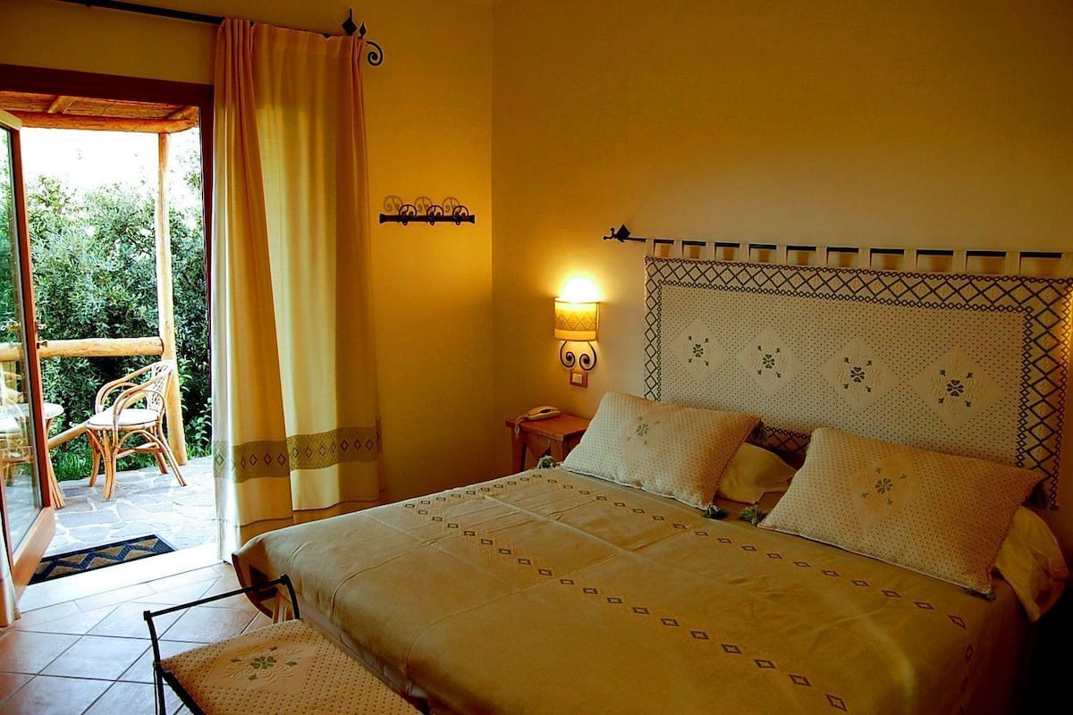 A bedroom with a bed, a nightstand, a wall-mounted lamp, a window with curtains, and a view of the outdoors.