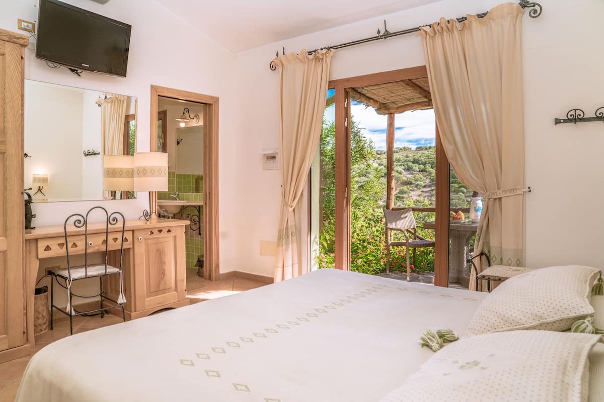 A bedroom with a bed, a vanity with a mirror, a television, and a view of greenery outside.