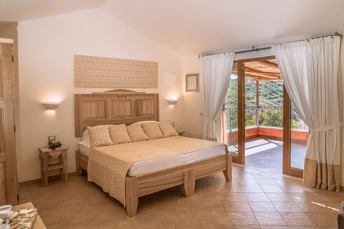 A bedroom with a wooden bed, a nightstand with a telephone, a wall-mounted light, and a view of a balcony with curtains.