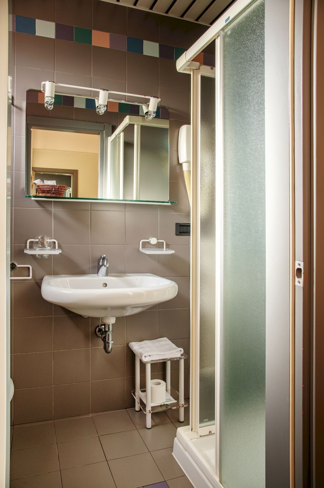 A bathroom with a white sink, a small stool with a towel, and a glass shower enclosure.
