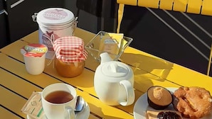 Una mesa amarilla dispuesta con una taza de té, una tetera, un tarro de mermelada, un plato con tostadas y mermelada, y un pastel en un balcón con vistas a edificios.