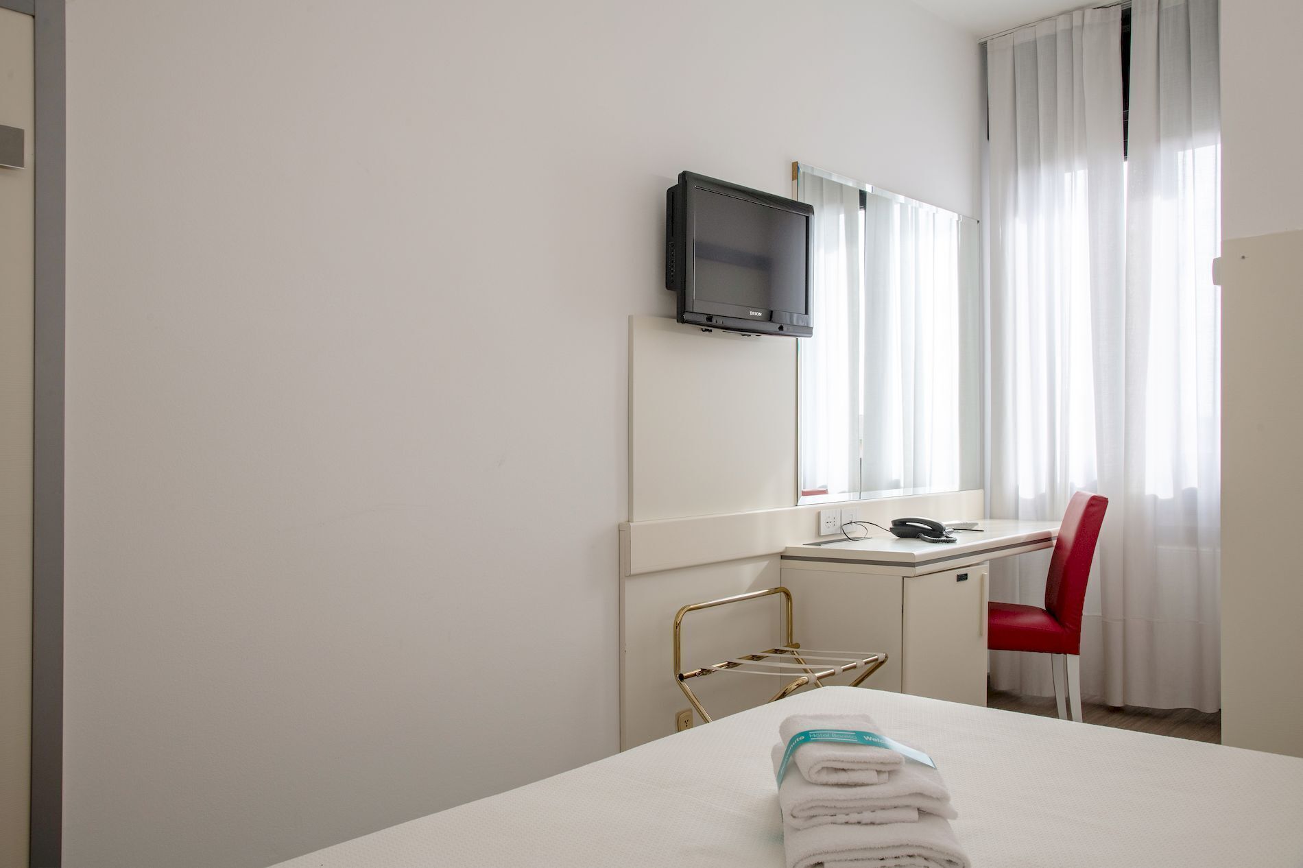 Ein Hotelzimmer mit Bett, Schreibtisch, Sessel und einem an der Wand montierten Fernseher. Ein Fenster mit Vorhängen.