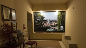 Habitación de hotel con un ventanal grande que ofrece vistas a un paisaje urbano, una silla, una mesita con un florero y cuadros en la pared.
