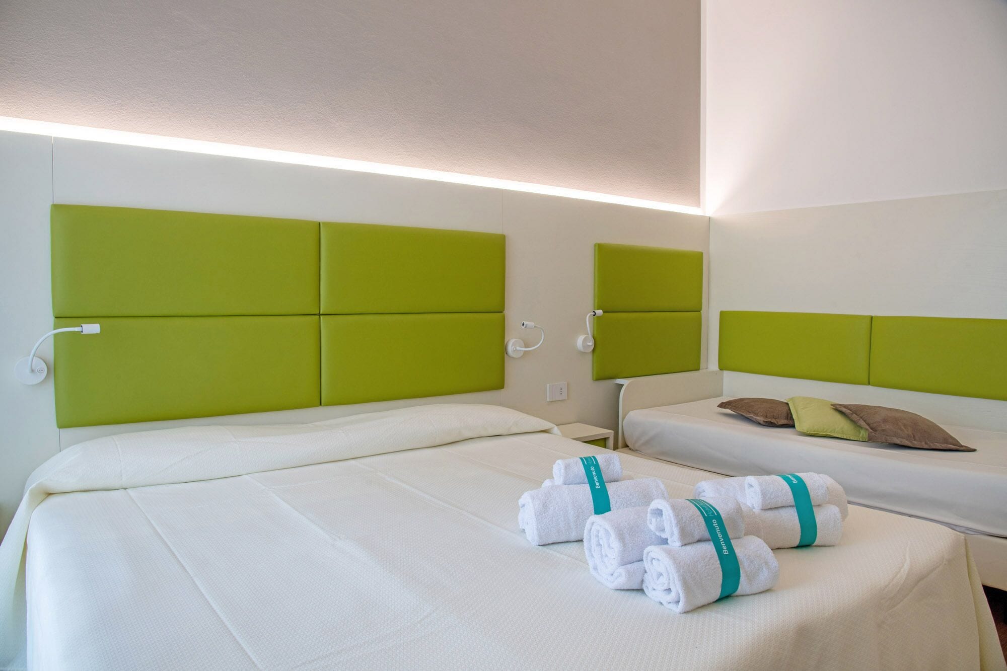A hotel room with two beds, each with a green headboard, white bedding, and a pair of rolled towels with a green ribbon.
