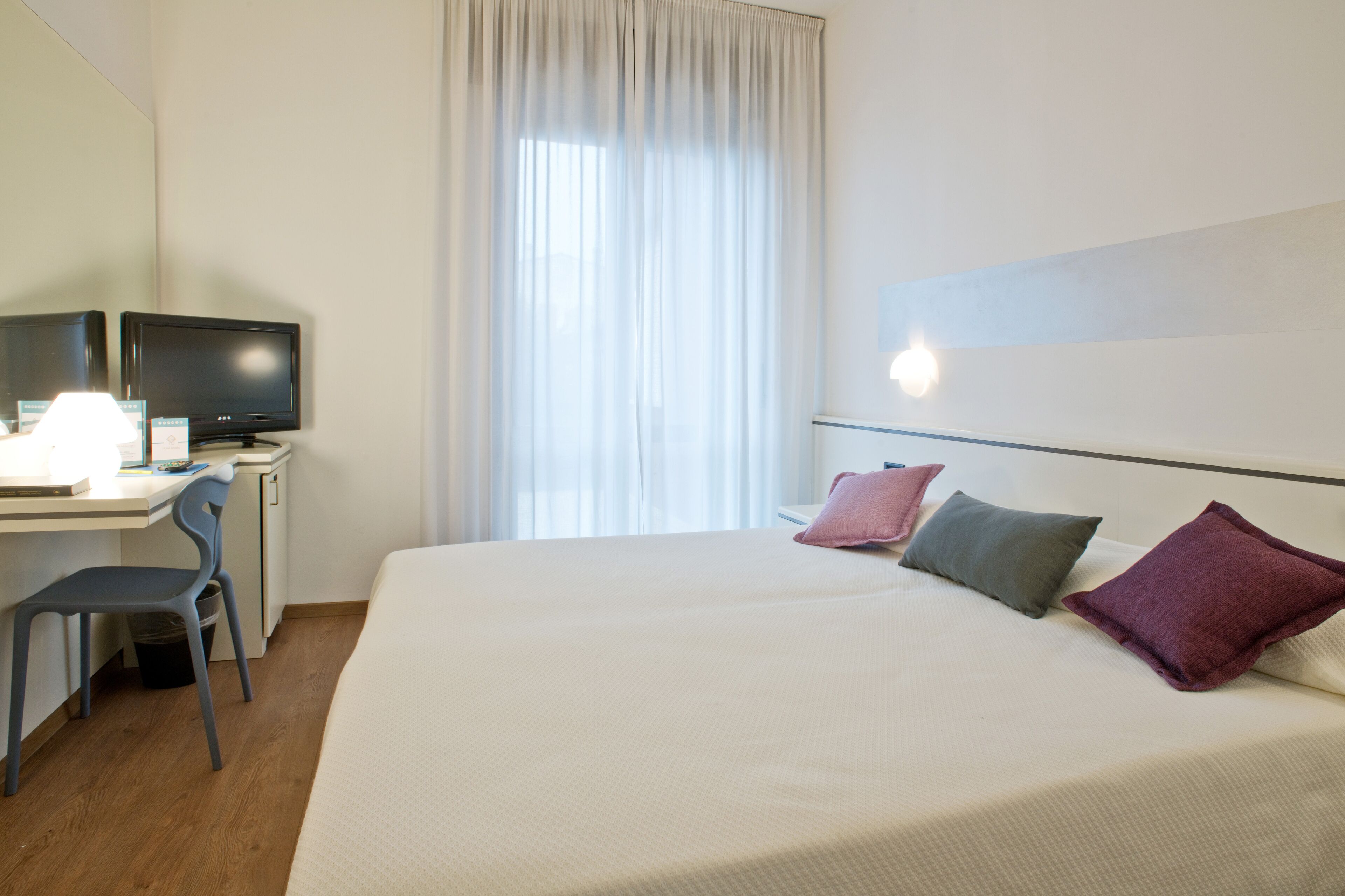 A hotel room with a bed, a desk with a computer, a chair, and a window with curtains.