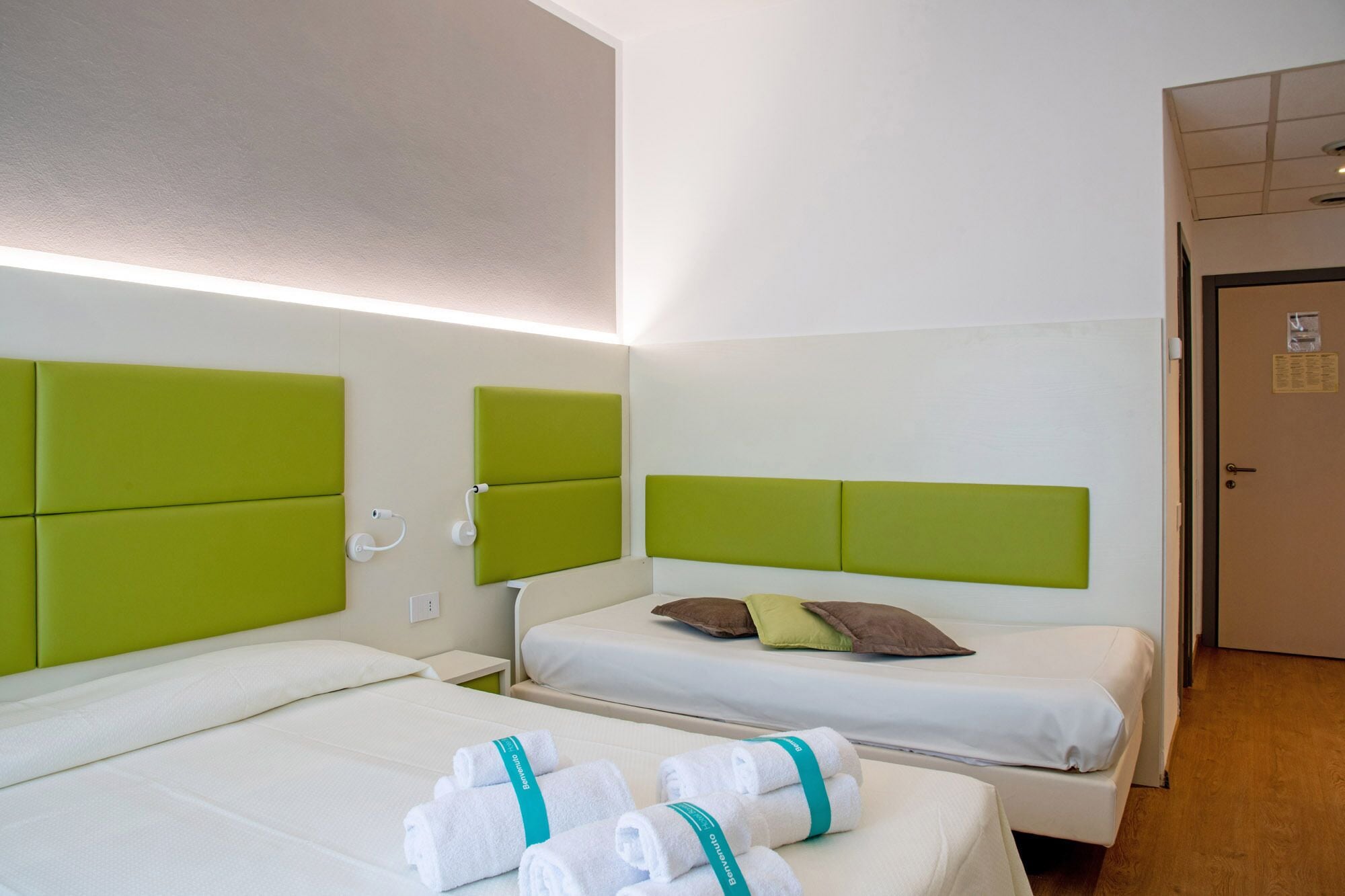 Una camera d'albergo con due letti, ciascuno con una testiera verde, biancheria bianca e un paio di asciugamani con un nastro color acquamarina.