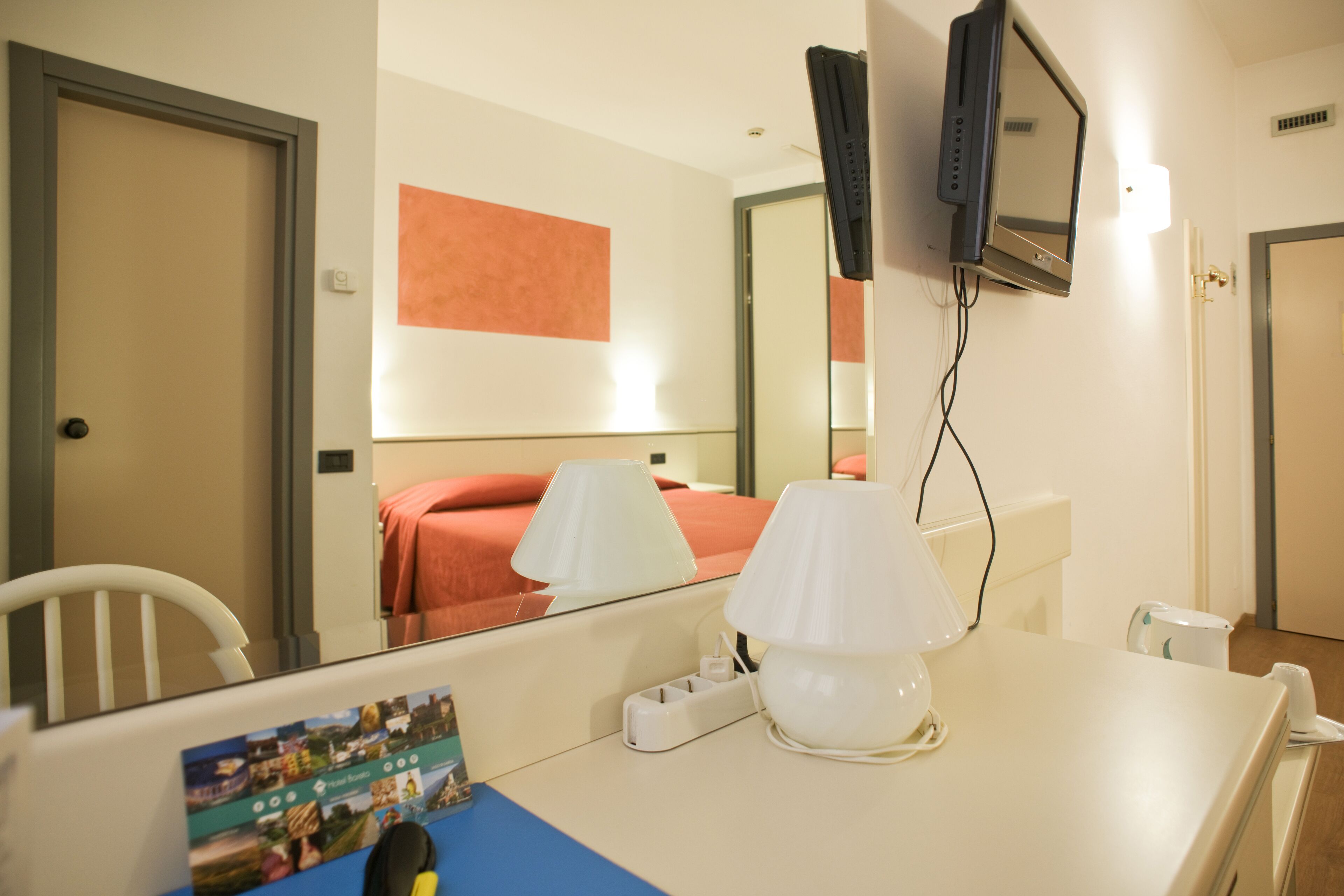 A hotel room with a bed, a desk with a lamp, a television mounted on the wall, and a mirror.