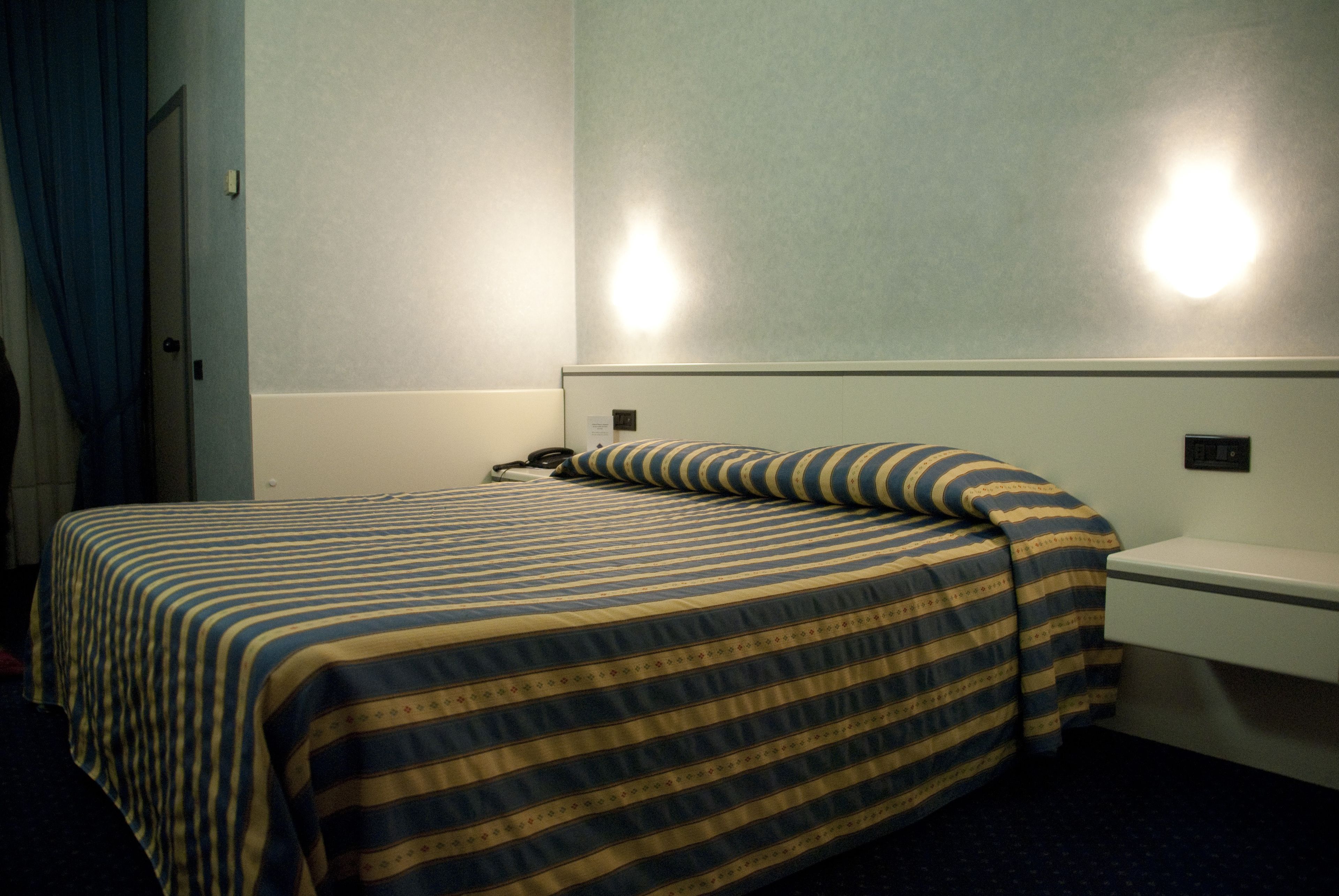 Quarto de hotel com cama, mesa de cabeceira e luminária fixada na parede.