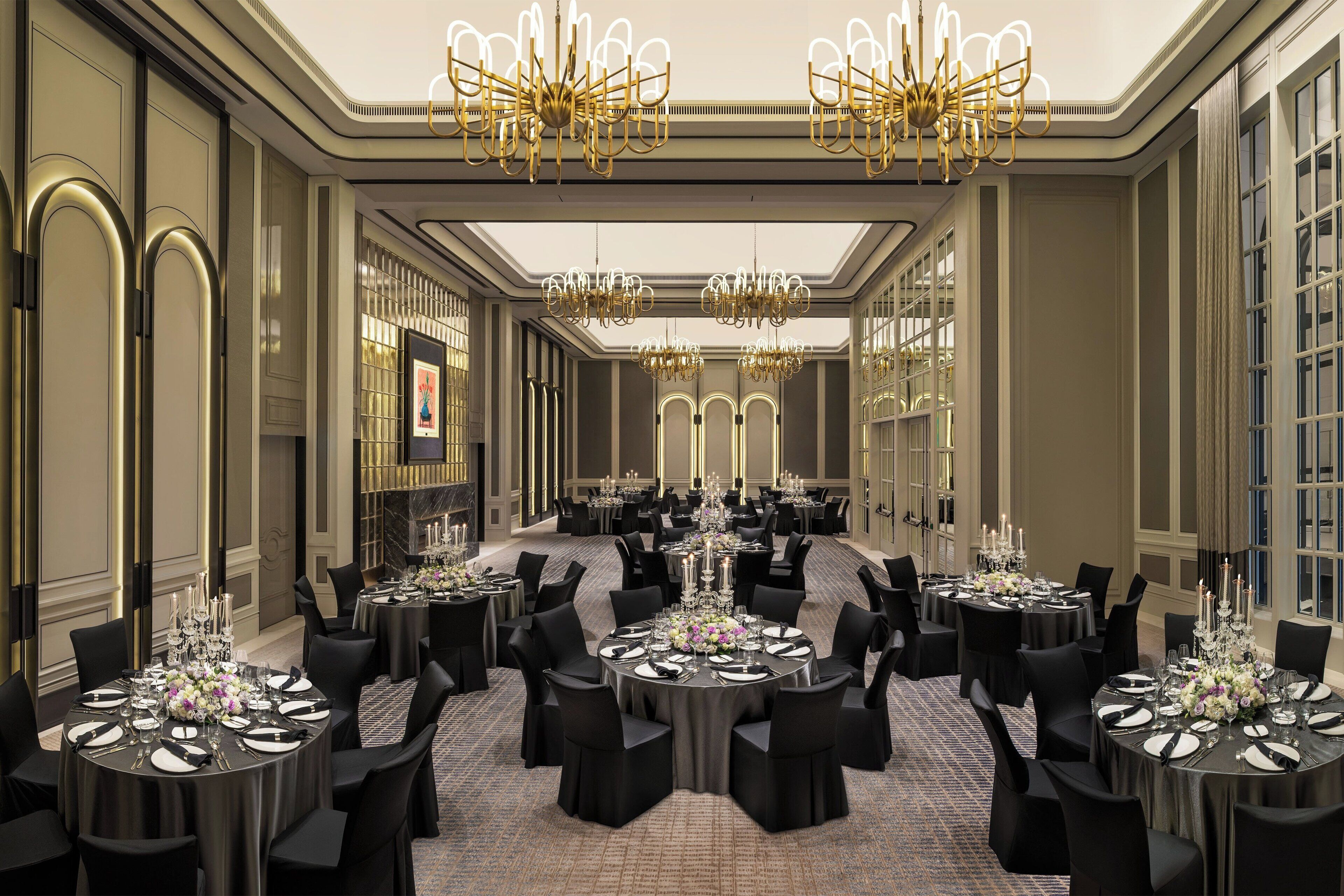 A large banquet hall with multiple round tables, black chairs, and chandeliers.