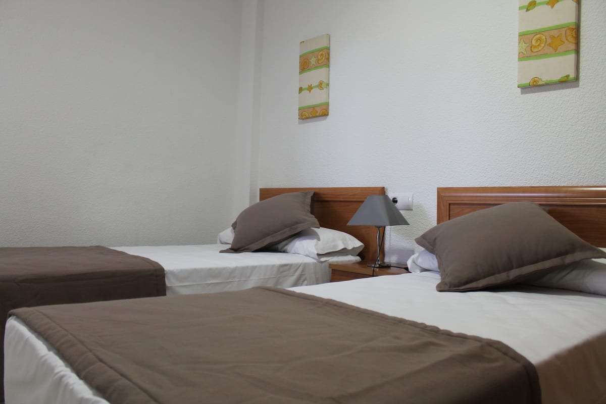 A hotel room with two single beds, each with a brown headboard and pillow, a bedside table with a lamp, and a wall-mounted artwork.