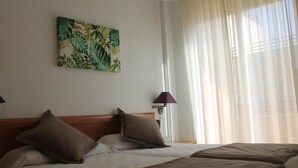 Una habitación de hotel con una cama, dos almohadas, una mesita de noche con una lámpara y un cuadro con motivos tropicales en la pared.