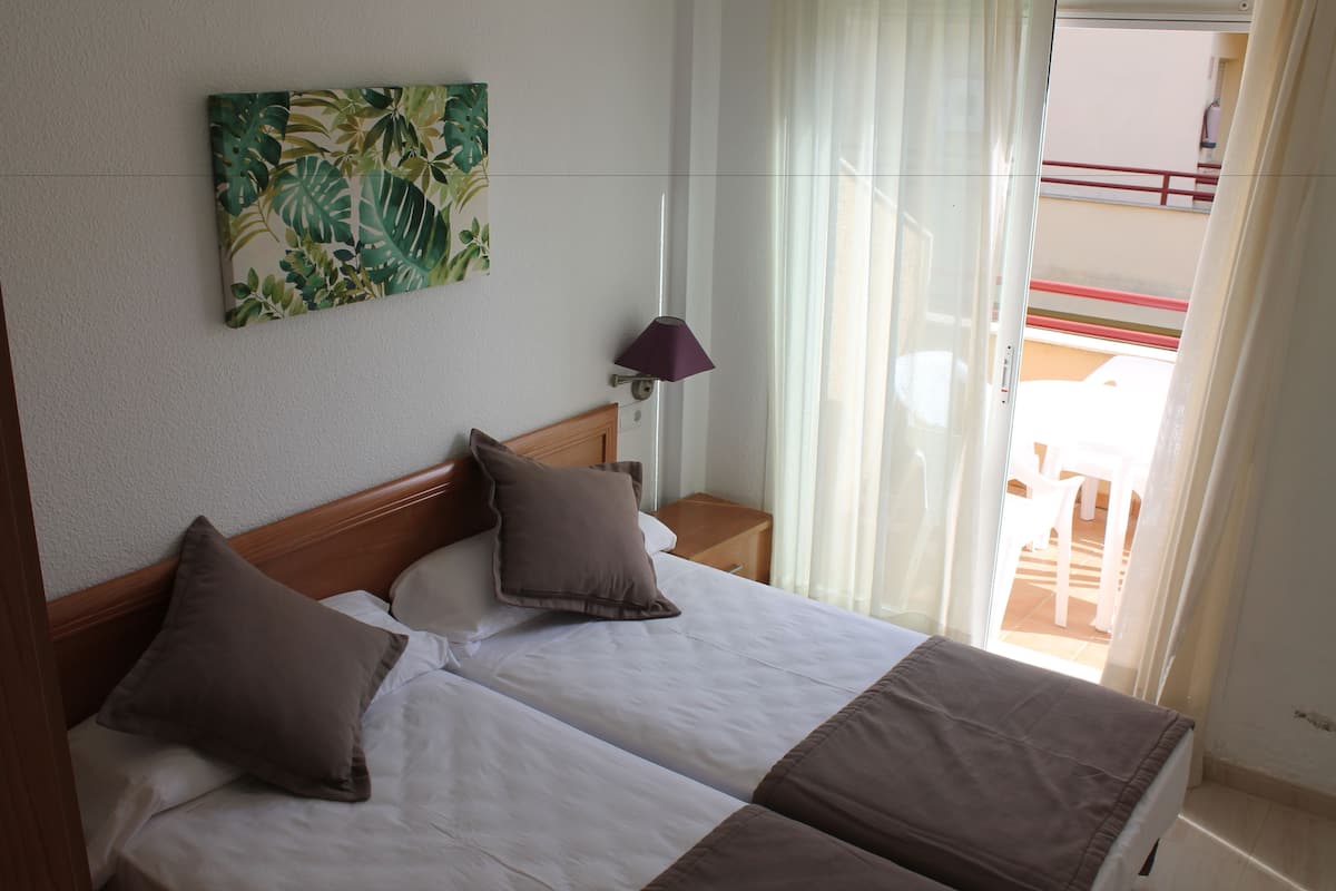 A hotel room with two beds, a nightstand, a lamp, a window with curtains, and a balcony with a railing.