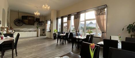 A dining area with tables set for two, a kitchen in the background, and large windows with curtains.