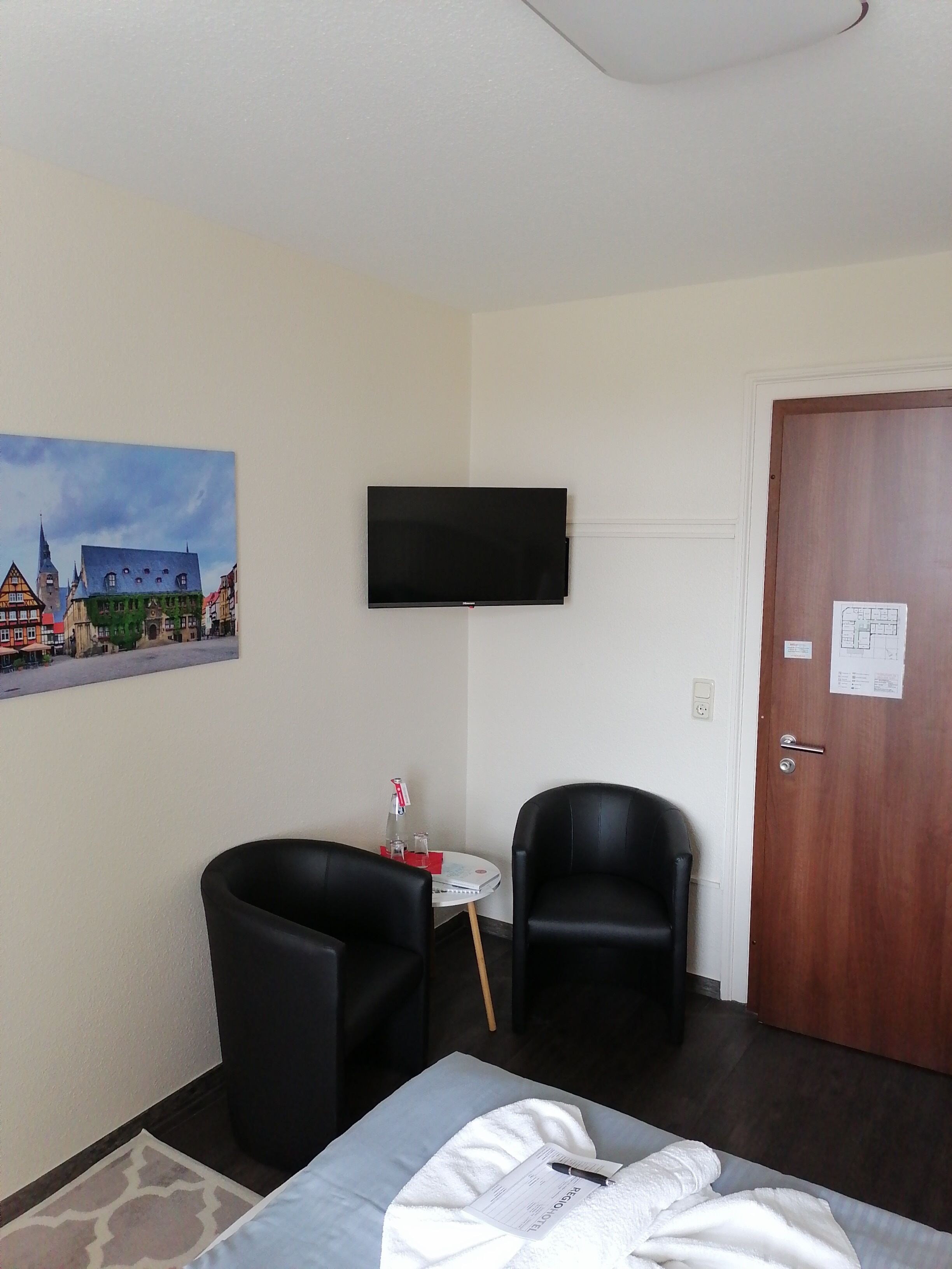 A hotel room with a bed, two black chairs, a small round table with a bottle and glasses, a painting of a town, a flat-screen TV, and a wooden door.