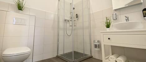 A modern bathroom with a glass-enclosed shower, a white toilet, a sink with a mirror, and a shelf with towels.