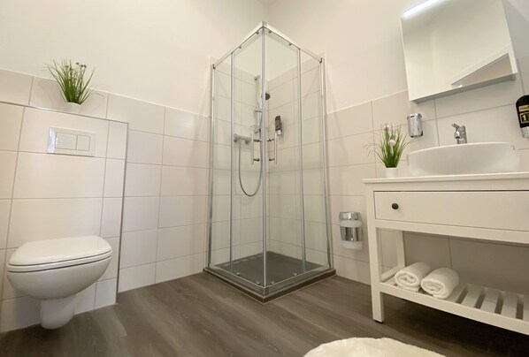 Une salle de bain moderne avec une douche à parois de verre, des toilettes blanches, un lavabo avec un miroir et une étagère avec des serviettes.