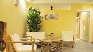 A room with a yellow wall, a large potted plant, a sofa, two chairs, a small table, and a picture on the wall.