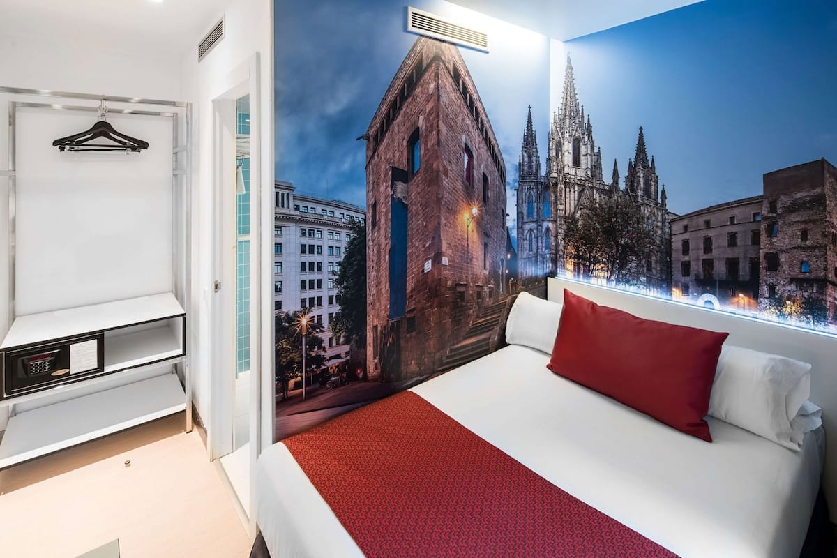 A hotel room with a bed, a hanging coat rack, a built-in TV, and a cityscape mural on the wall.