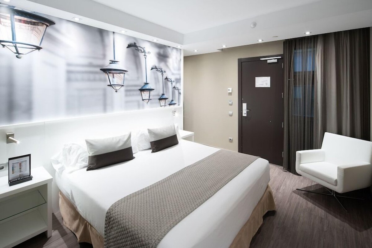 Une chambre d’hôtel avec un grand lit, une table de chevet avec une lampe, une chaise et une porte avec un rideau.