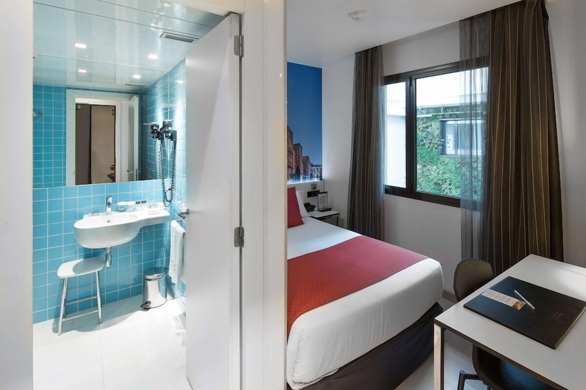 A hotel room with a bed, a desk, a bathroom with a sink and mirror, and a window with curtains.