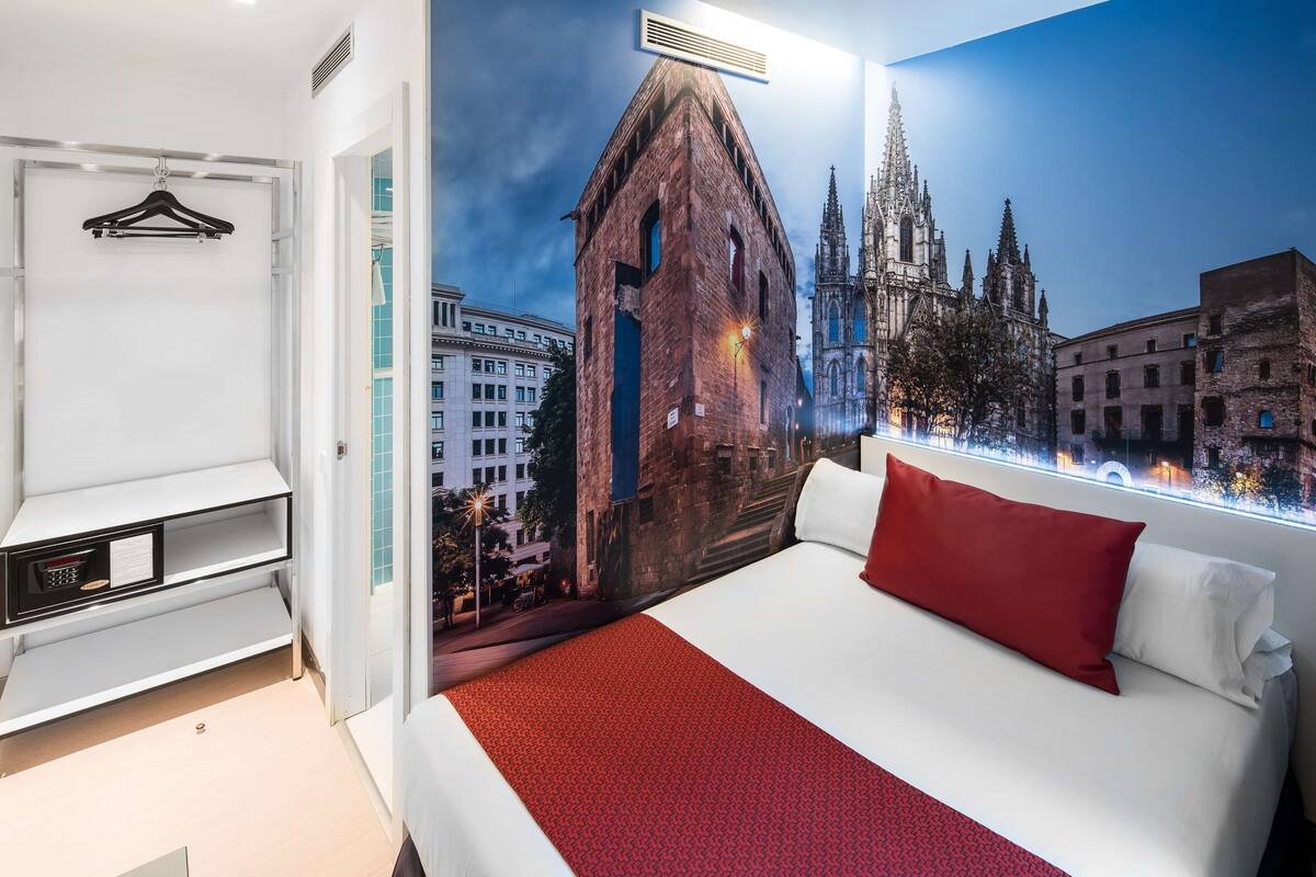 Een hotelkamer met een groot bed, een rood kussen, een wit kussen, een wandkapstok en een muurschildering van een gotische kathedraal.