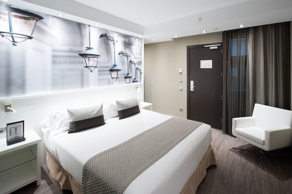 Een moderne hotelkamer met een groot bed, een witte fauteuil, een nachtkastje met een lamp, en uitzicht op de stad door het raam.