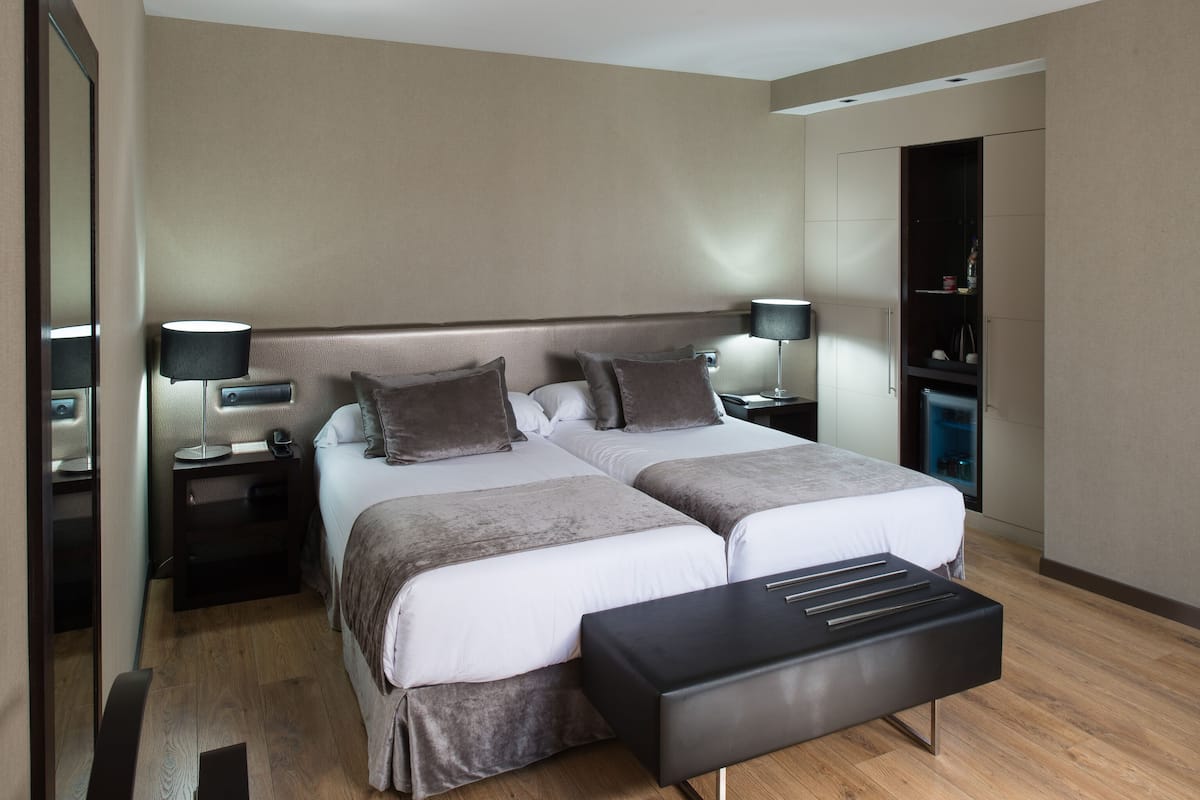 Een moderne hotelkamer met twee bedden, een nachtkastje met een lamp, een kledingkast en een bankje.
