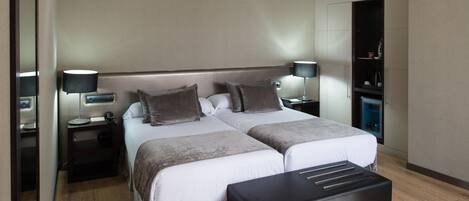 A modern hotel room with two beds, a bedside table with a lamp, a wardrobe, and a bench.