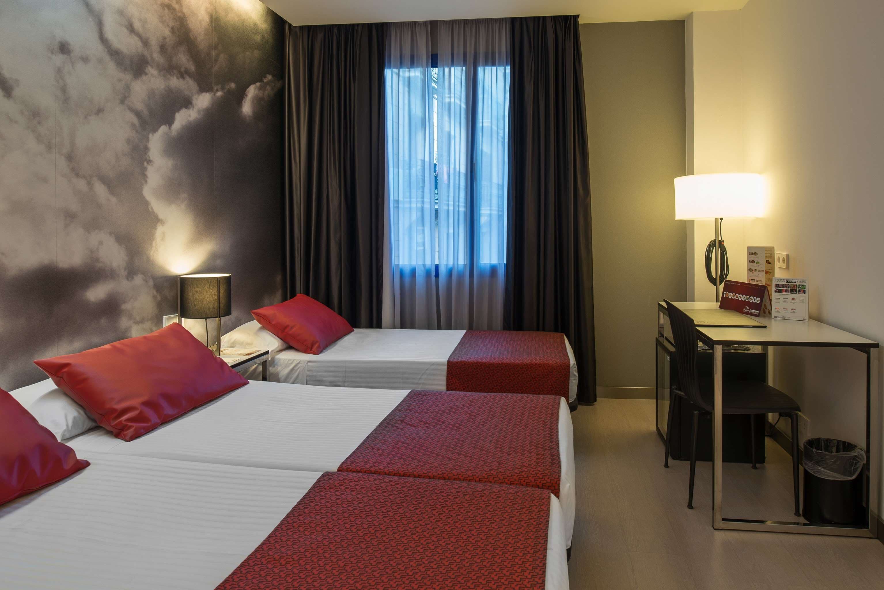 Een hotelkamer met twee bedden, een bureau, een stoel, een lamp en een raam met gordijnen.
