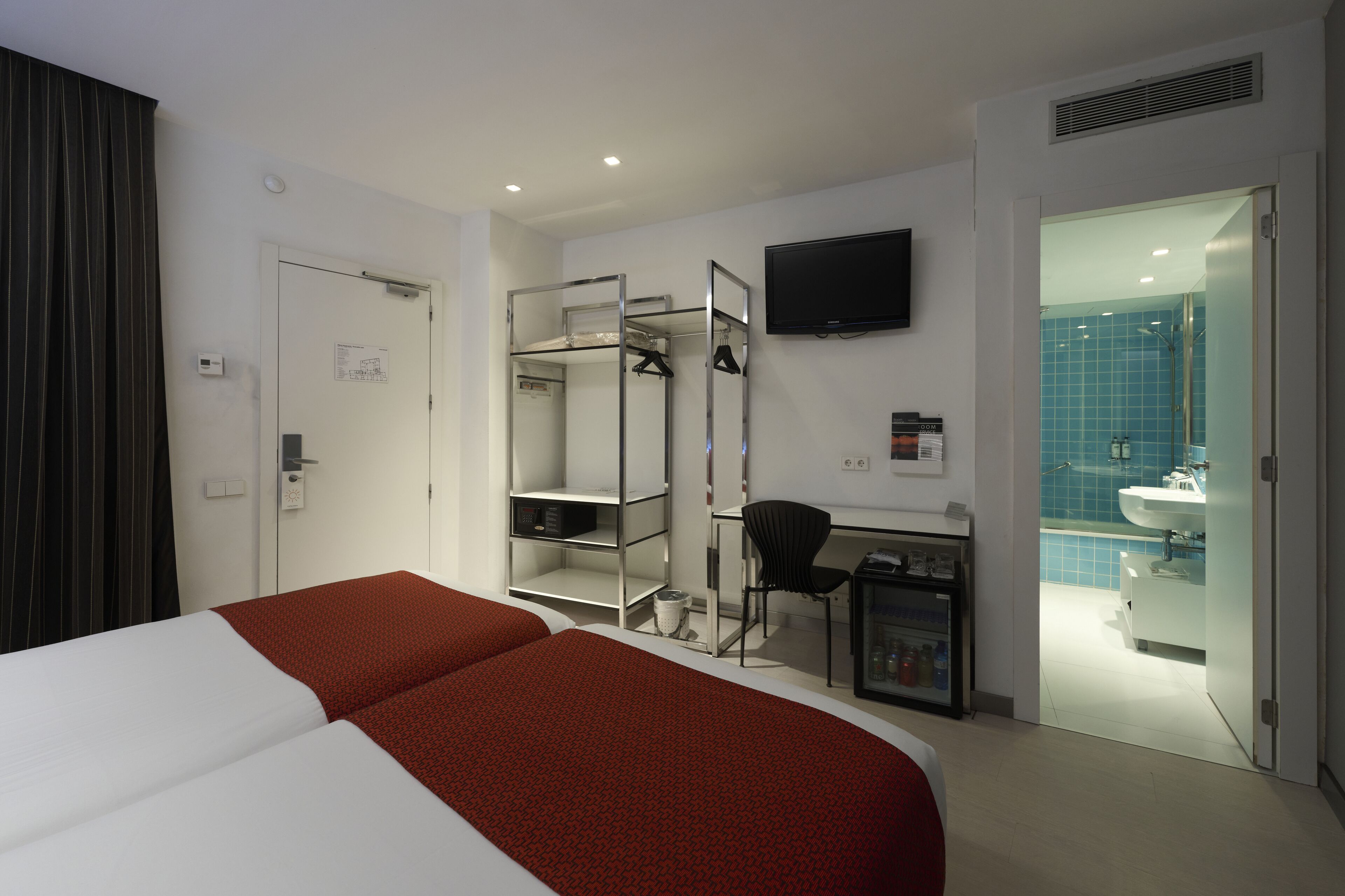 A hotel room with a bed, a desk, a chair, a TV, and a bathroom visible through an open door.