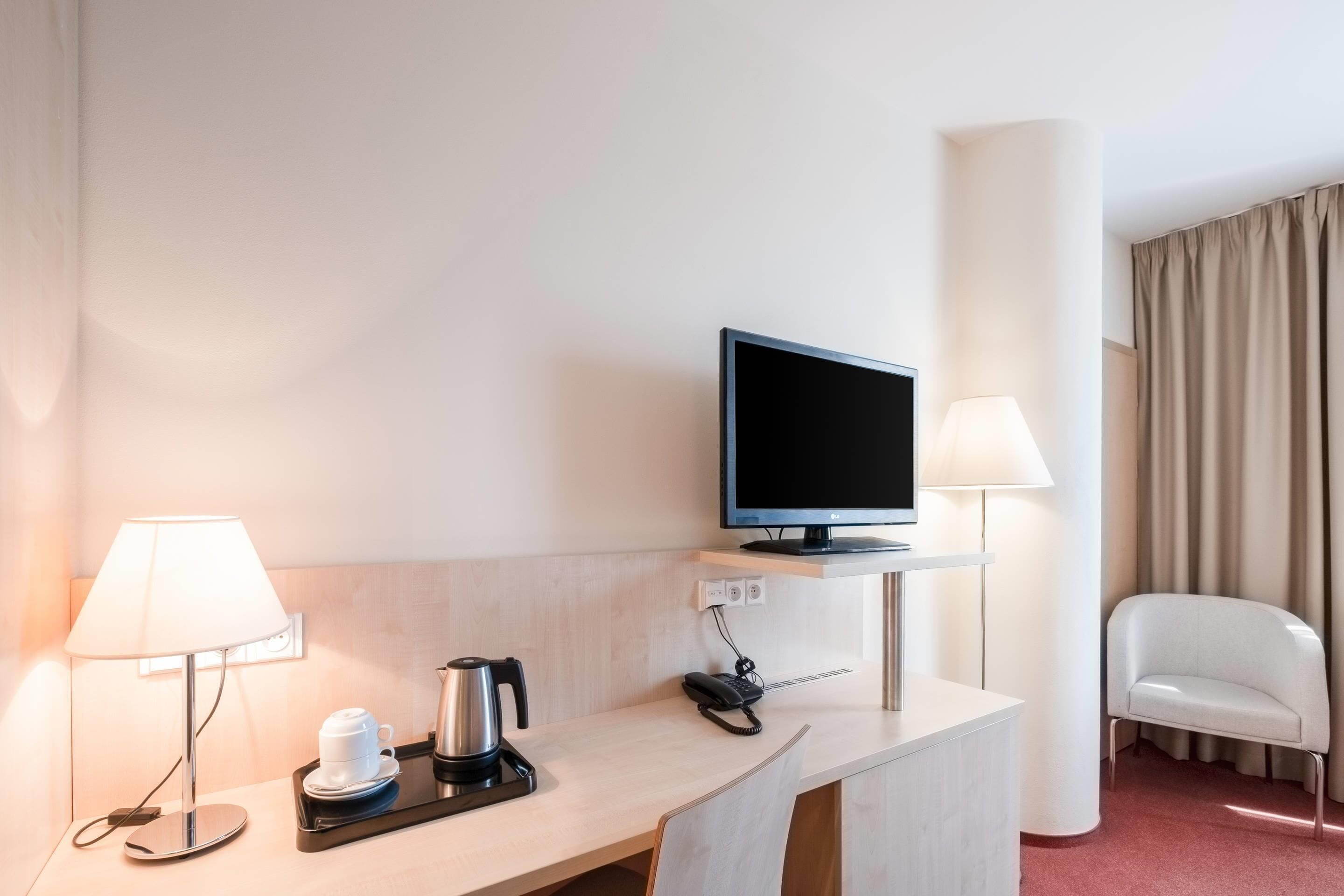 A hotel room with a desk, a television, a lamp, a chair, and a coffee maker.