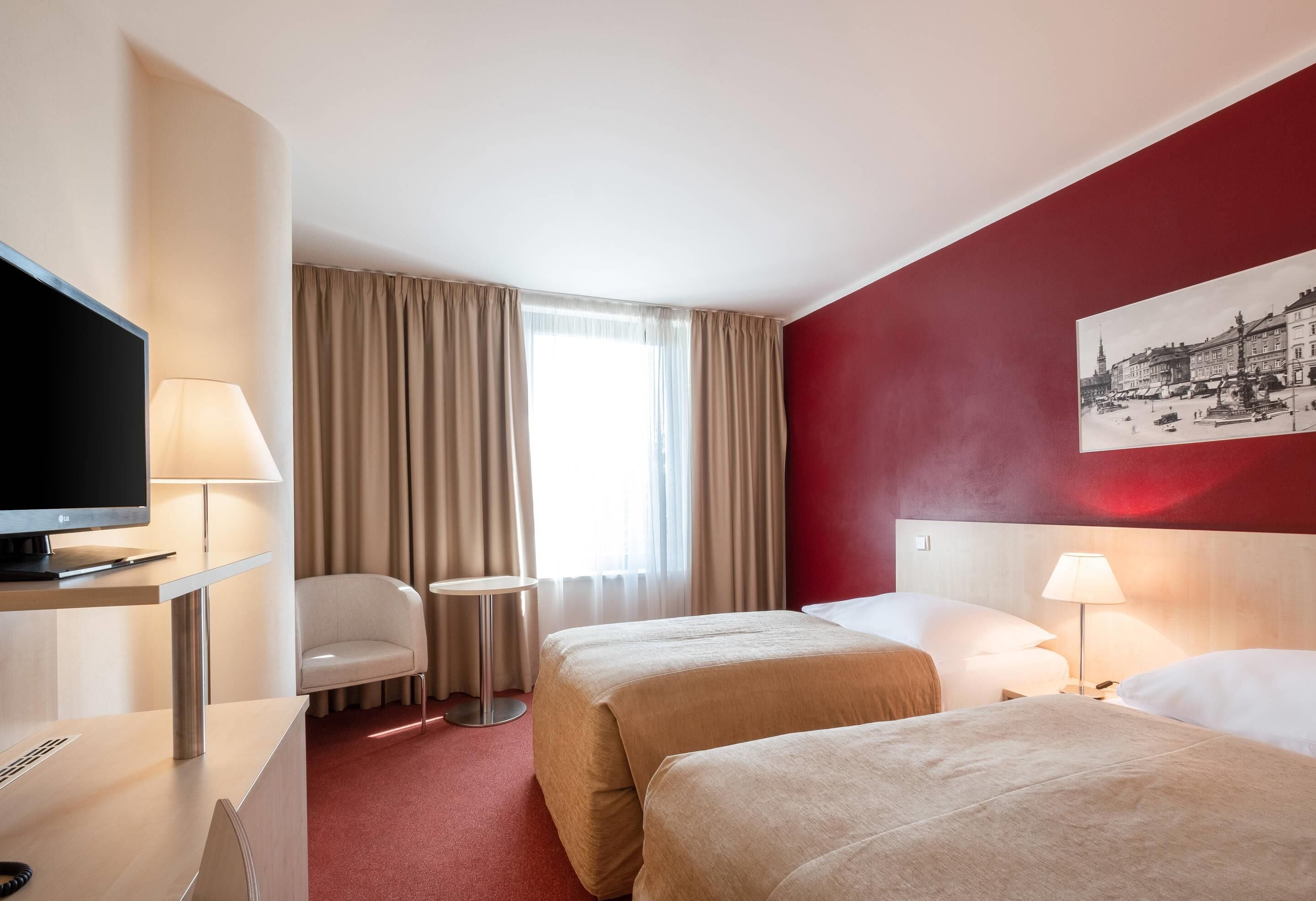 A hotel room with two beds, a desk with a TV, a chair, a small table, and a window with curtains.