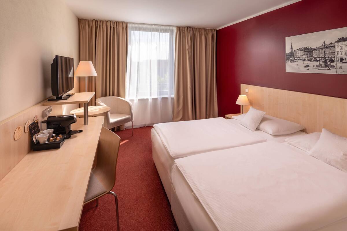 Quarto de hotel com escrivaninha, duas camas, uma cadeira, televisão e janela com cortinas.
