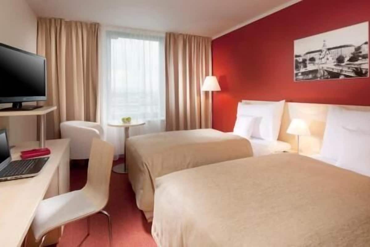 Quarto de hotel com duas camas, uma escrivaninha com um laptop, uma cadeira, uma televisão e uma pequena mesa com abajur.