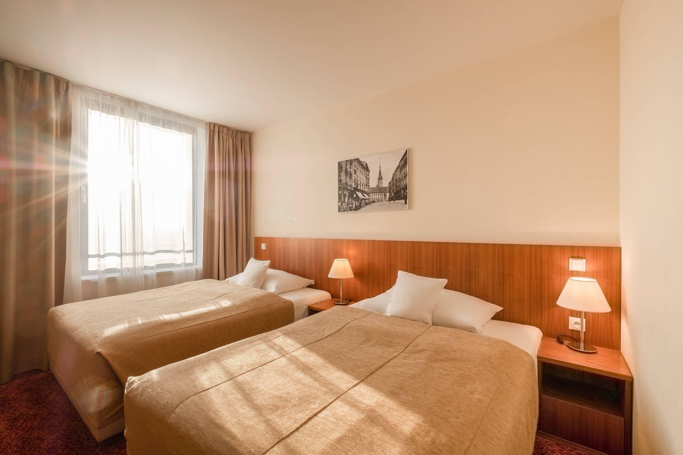 Une chambre d’hôtel avec deux lits, une tête de lit en bois, une table de chevet avec une lampe, un tableau encadré au mur et une grande fenêtre avec des rideaux.