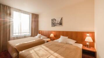 Une chambre d’hôtel avec deux lits, une tête de lit en bois, une table de chevet avec une lampe, un tableau encadré au mur et une grande fenêtre avec des rideaux.