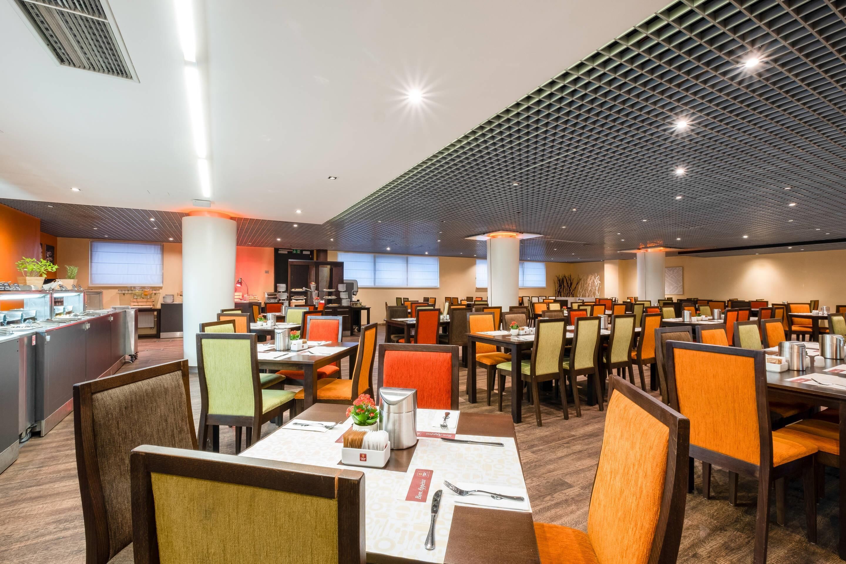 A spacious dining area with multiple tables and chairs, a kitchen area, and a ceiling with a grid pattern.