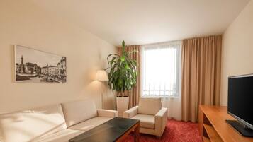 Habitación de hotel con sofá, dos sillones, una mesita de centro, televisor, ventana grande con cortinas, lámpara de pie, planta en maceta y cuadro de un paisaje urbano.