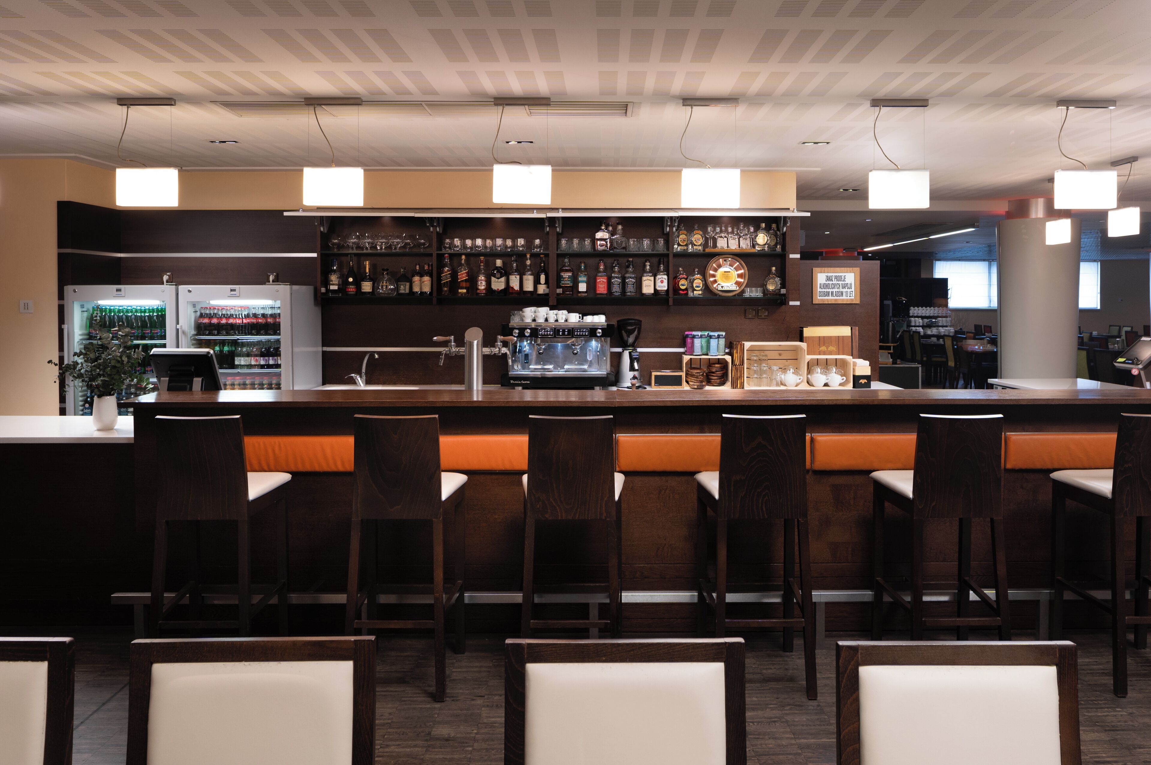 A bar area with a long counter, multiple bar stools, and a coffee machine. There are shelves with bottles and glasses behind the counter.