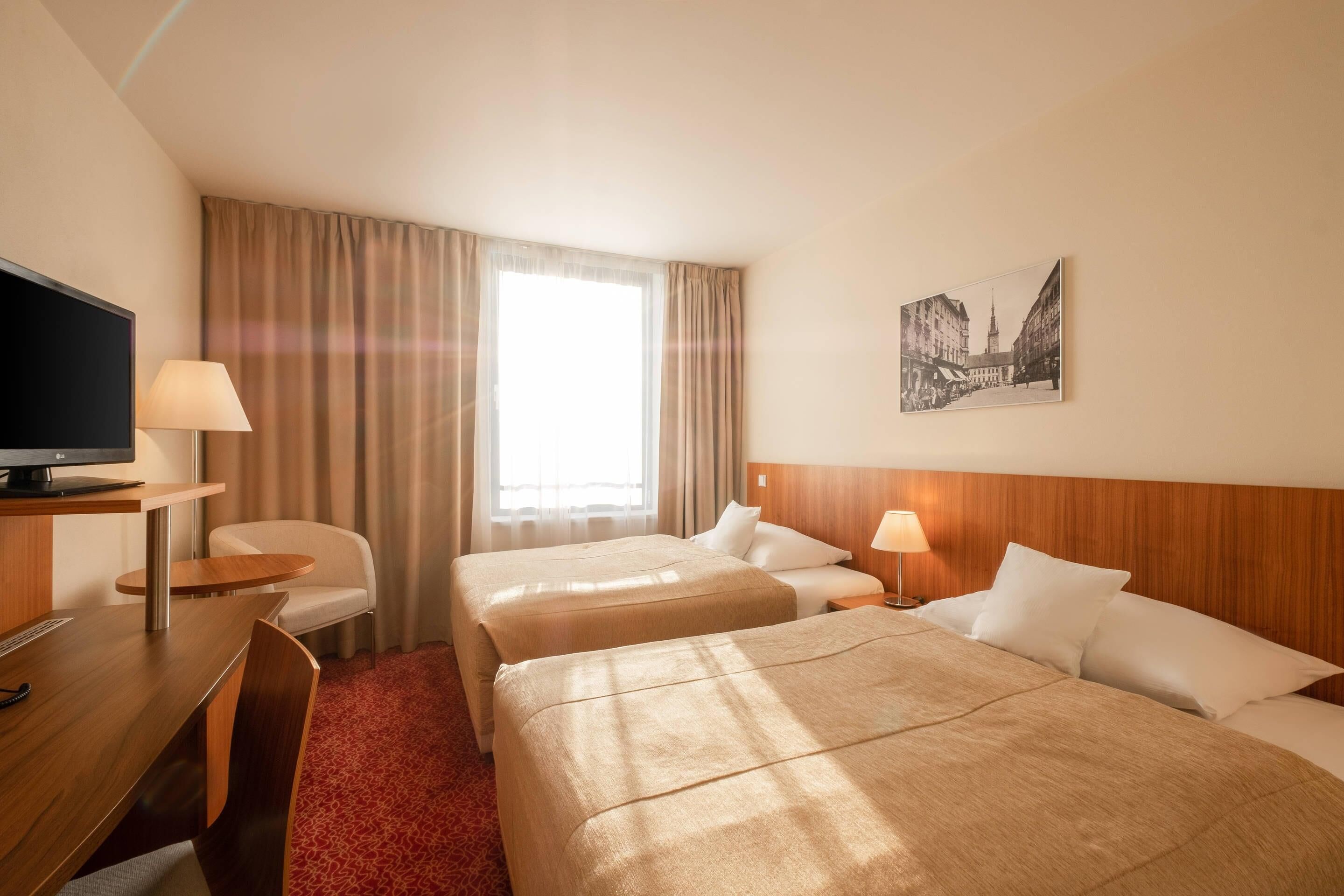 Een hotelkamer met twee bedden, een bureau met een televisie, een stoel, een lamp en een lijst aan de muur.