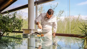 A chef pouring sauce into a dish on a reflective glass table in a modern kitchen with large windows. - Hotel Villa Rizzo Resort & Spa (San Cipriano Picentino)