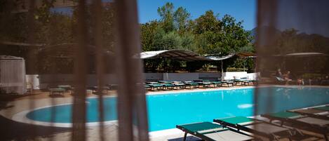 A pool area with lounge chairs and umbrellas, surrounded by trees and a clear sky.