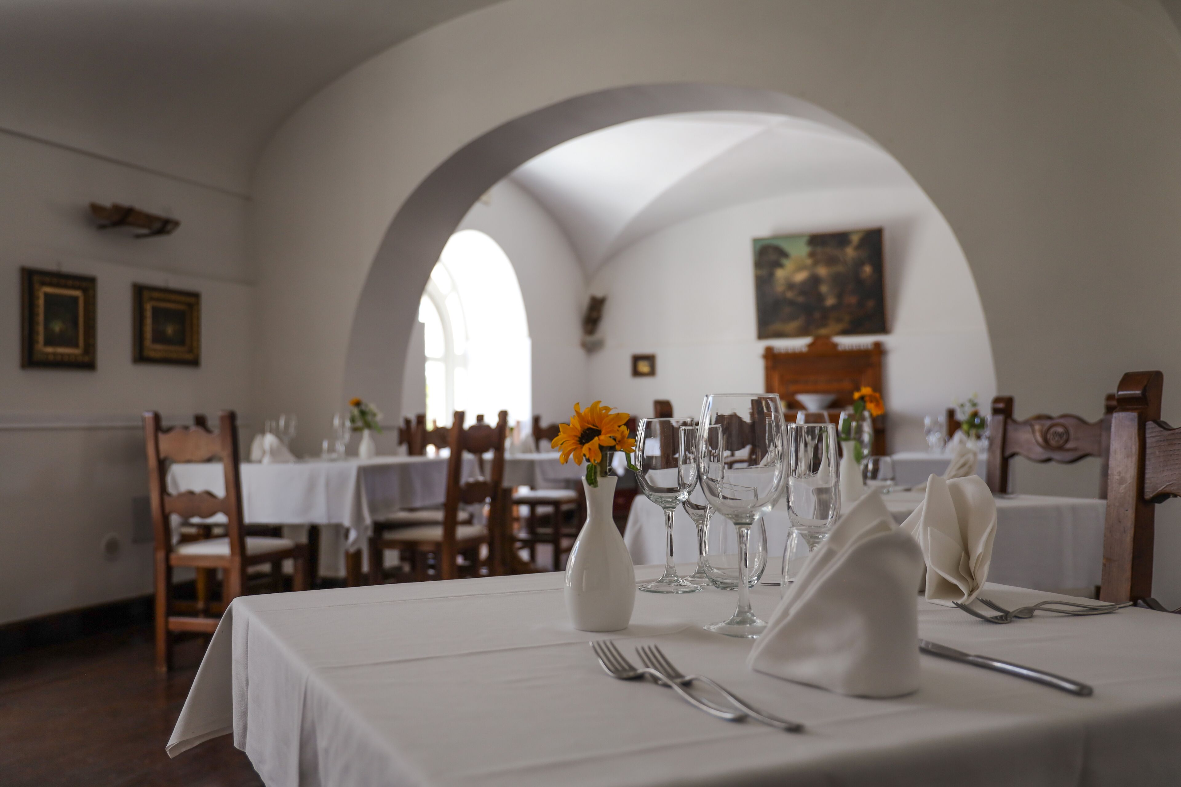 Uma sala de jantar com mesas postas para refeição, com toalhas de mesa brancas, copos de vinho e um vaso com flores.
