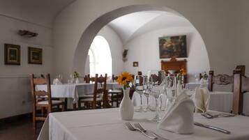 Uma sala de jantar com mesas postas para refeição, com toalhas de mesa brancas, copos de vinho e um vaso com flores.