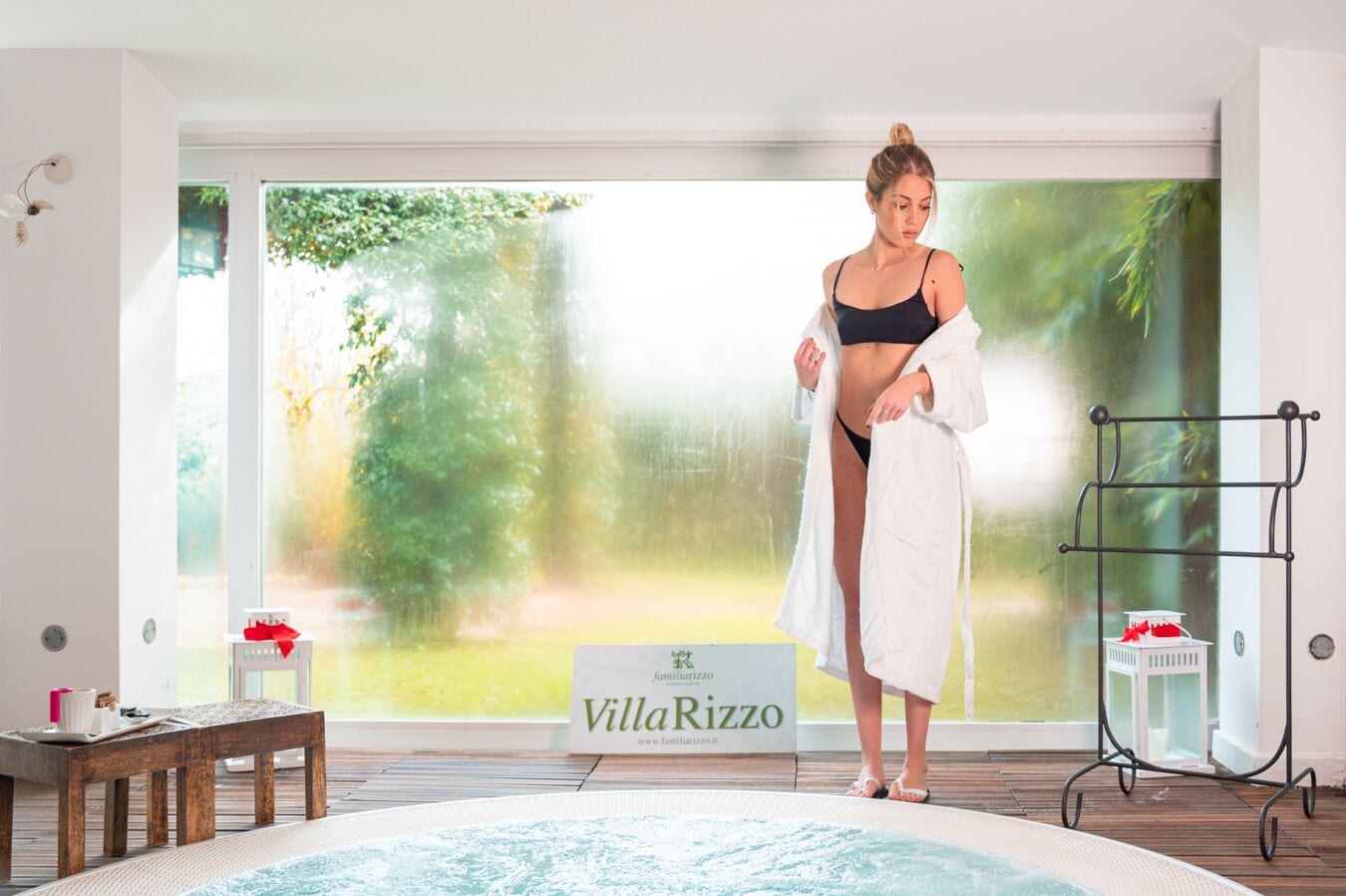 En kvinna i en svart bikini och en vit morgonrock står vid en pool, och i bakgrunden syns en skylt med texten ”Villa Rizzo”.
