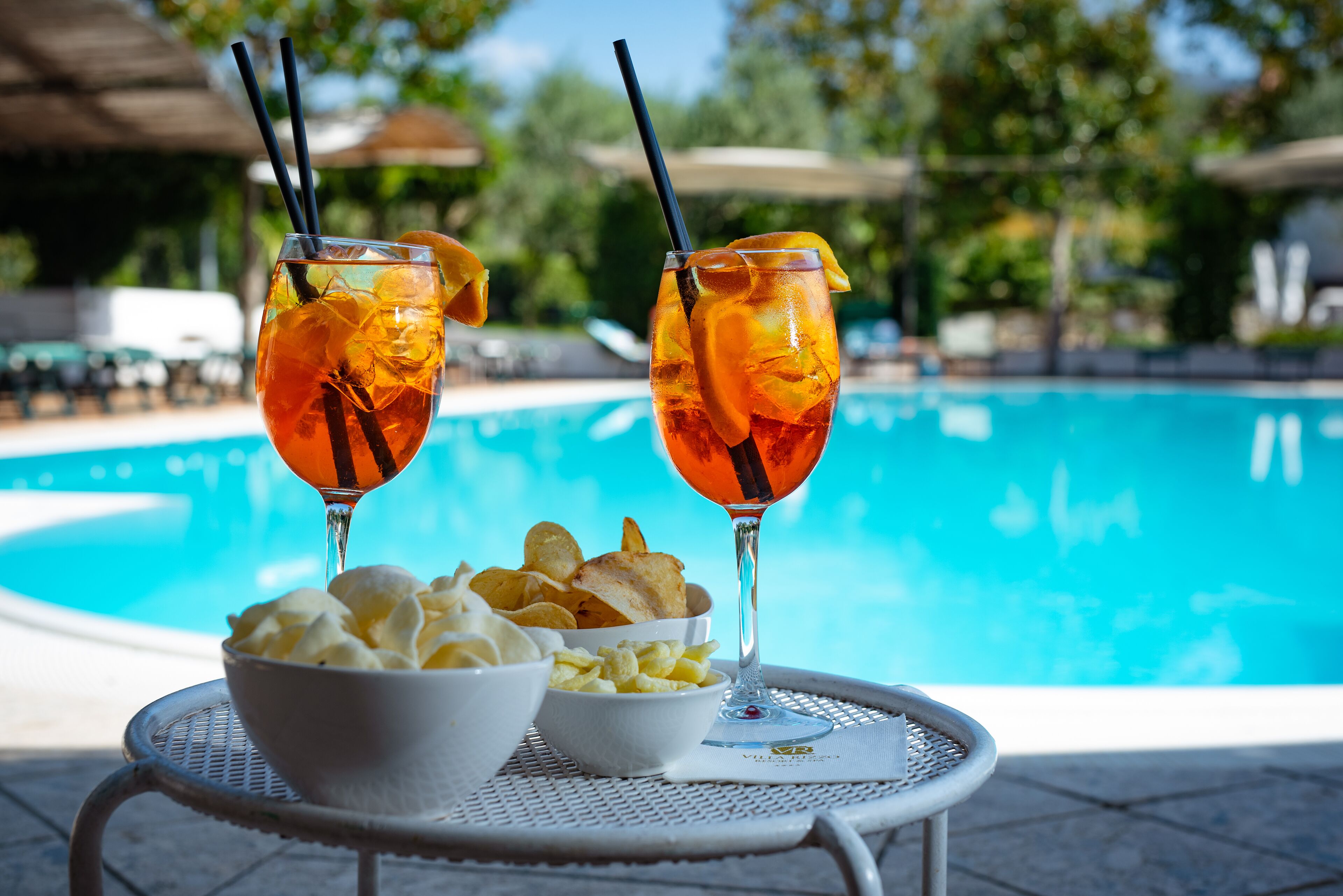 A poolside setting with two drinks in glasses, a bowl of chips, and a bowl of dip.