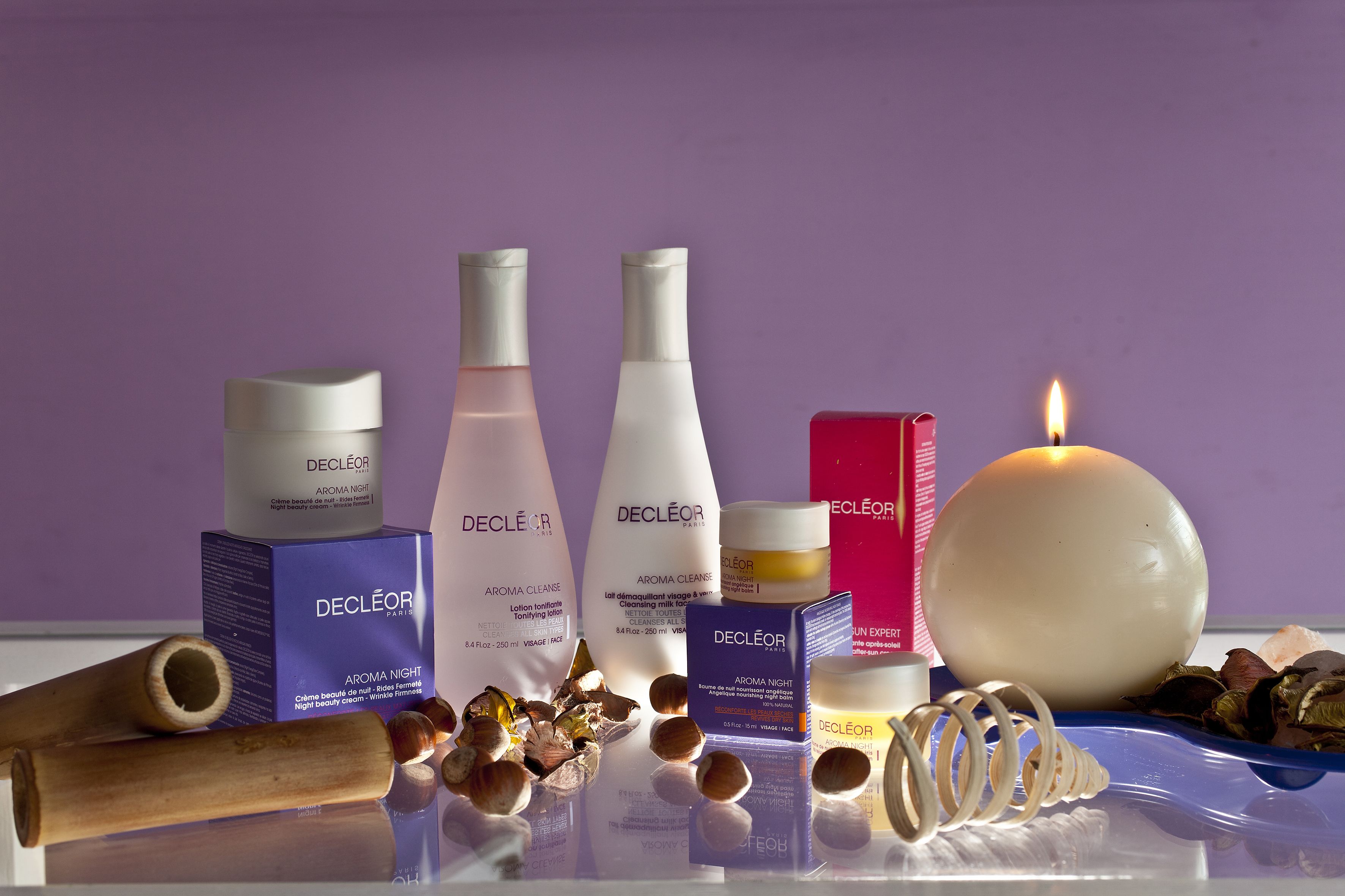 A selection of skincare products, including bottles and boxes, arranged on a reflective surface with a candle and natural elements like nuts and a wooden stick.